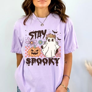 K&ouml;nnte beinhalten: Ein lavendelfarbenes T-Shirt mit einem Aufdruck eines Geistes, der eine Blumenkrone tr&auml;gt und ein Peace-Zeichen macht. Der Geist befindet sich neben einem K&uuml;rbis und der Text "Stay Spooky" ist unter dem Aufdruck gedruckt.