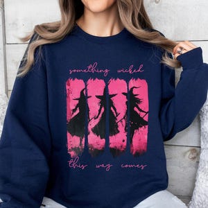 以下が含まれることがあります： ネイビーブルーのスウェットシャツ。ピンク色の背景に、3人の魔女のシルエットが描かれたピンク色のグラフィック。テキスト「something wicked this way comes」がグラフィックの上と下にあります。クルーネックと長袖です。