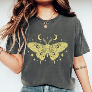 以下が含まれることがあります： 黄色い蛾のデザイン、三日月と星が描かれた濃いグレーのTシャツ。「Pepper」の文字が下部にプリントされています。カジュアルなスタイルです。