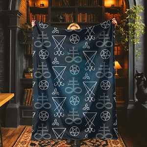 May include: A dark blue blanket with a repeating white pattern of sigils, pentagrams, and other occult symbols. The blanket is held up in front of a person, with a bookshelf and a black cat in the background.
