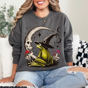 Puede incluir: Una sudadera gris carbón con un gráfico de una rana verde con un sombrero de bruja sentada en una luna creciente con una calavera. La rana está rodeada de setas y estrellas.