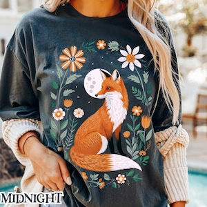 以下が含まれることがあります： 濃いグレーのTシャツに、花畑に座っているキツネと満月のグラフィックがプリントされています。キツネはオレンジと白で、花はピンク、イエロー、ホワイトの色合いです。
