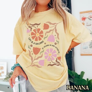 Pu&ograve; includere: Una maglietta gialla con un disegno floreale e il testo "Life is a balance of holding on and letting go".