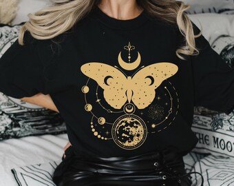 以下が含まれることがあります： 金色の蝶の模様、月の満ち欠け、星が描かれた黒いTシャツ。デザインはシャツの中央にあり、三日月と三日月の上に星が描かれています。