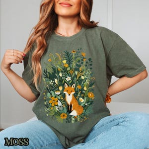 May include: A green t-shirt with a graphic of a fox sitting in a field of flowers. The fox is orange and white with a bushy tail. The flowers are a variety of colors, including pink, yellow, and purple.