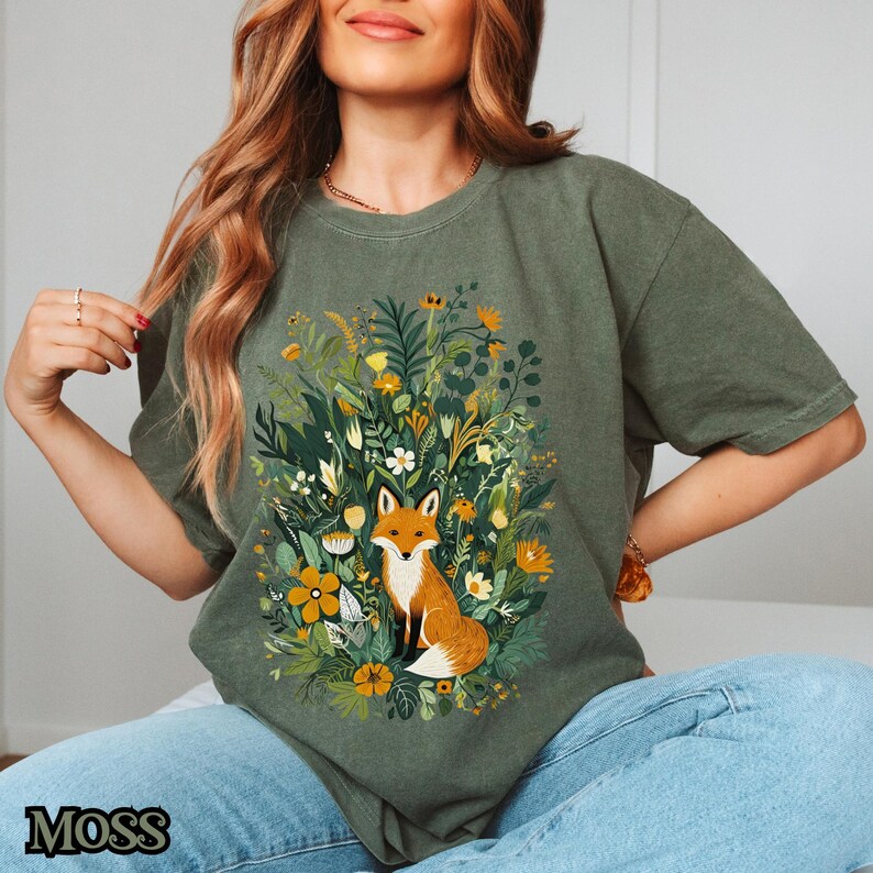 May include: A green t-shirt with a graphic of a fox sitting in a field of flowers. The fox is orange and white with a bushy tail. The flowers are a variety of colors, including pink, yellow, and purple.