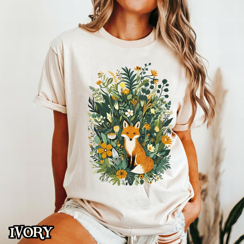 May include: A cream colored t-shirt with a graphic of a fox sitting in a field of flowers. The flowers are in shades of yellow, green, and white.
