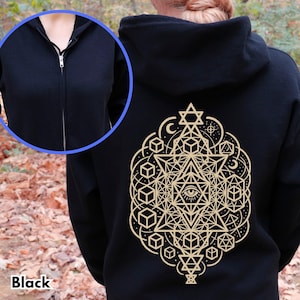 Puede incluir: Sudadera negra con cremallera y diseño geométrico beige en la espalda. El diseño incluye estrellas, cubos y un ojo. La sudadera tiene capucha y cremallera completa. La palabra "Black" está impresa en la parte inferior.