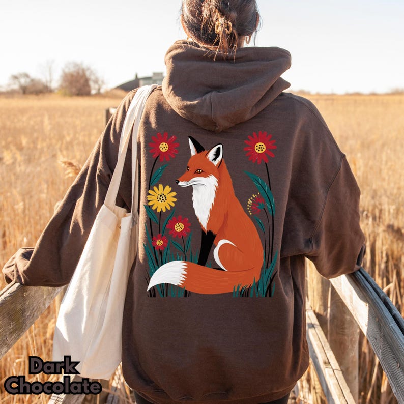May include: Dark chocolate brown hoodie featuring a vibrant illustration of a fox amidst colorful flowers. The fox is depicted in shades of orange, white, and black. The hoodie has a drawstring hood and a relaxed fit. The text "Dark Chocolate" is at the bottom.