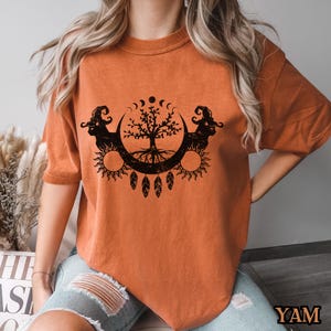 May include: A burnt orange t-shirt with a black graphic of a tree with roots, surrounded by a crescent moon and sun. The graphic also includes feathers and a crescent moon with stars.