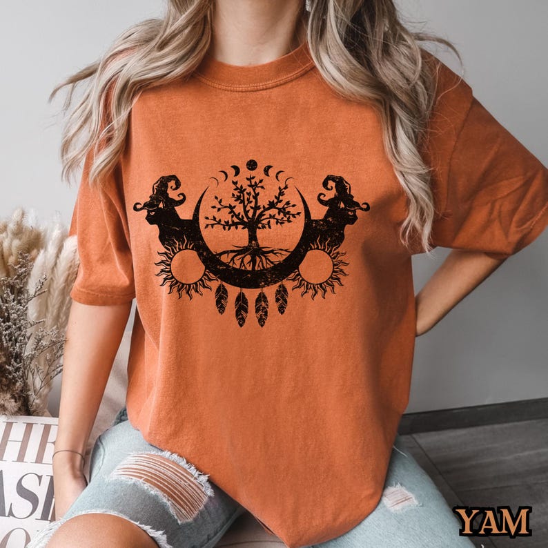 May include: A burnt orange t-shirt with a black graphic of a tree with roots, surrounded by a crescent moon and sun. The graphic also includes feathers and a crescent moon with stars.