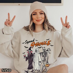 Pu&ograve; includere: Una felpa beige con il testo "Squad Ghouls" in arancione e nero. La grafica presenta uno scheletro, un fantasma e una strega che tiene un bicchiere di vino.
