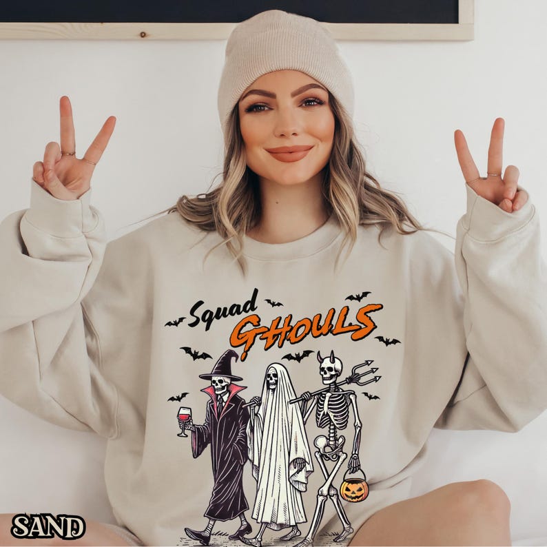 Pu&ograve; includere: Una felpa beige con il testo "Squad Ghouls" in arancione e nero. La grafica presenta uno scheletro, un fantasma e una strega che tiene un bicchiere di vino.