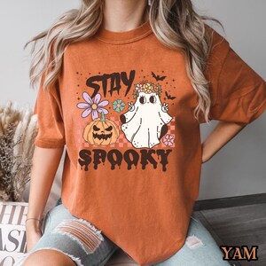 K&ouml;nnte beinhalten: Orangefarbenes T-Shirt mit einem Geist, der eine Blumenkrone tr&auml;gt und ein Peace-Zeichen macht. Der Text "Stay Spooky" ist mit einem K&uuml;rbis und Blumen auf das Shirt gedruckt.