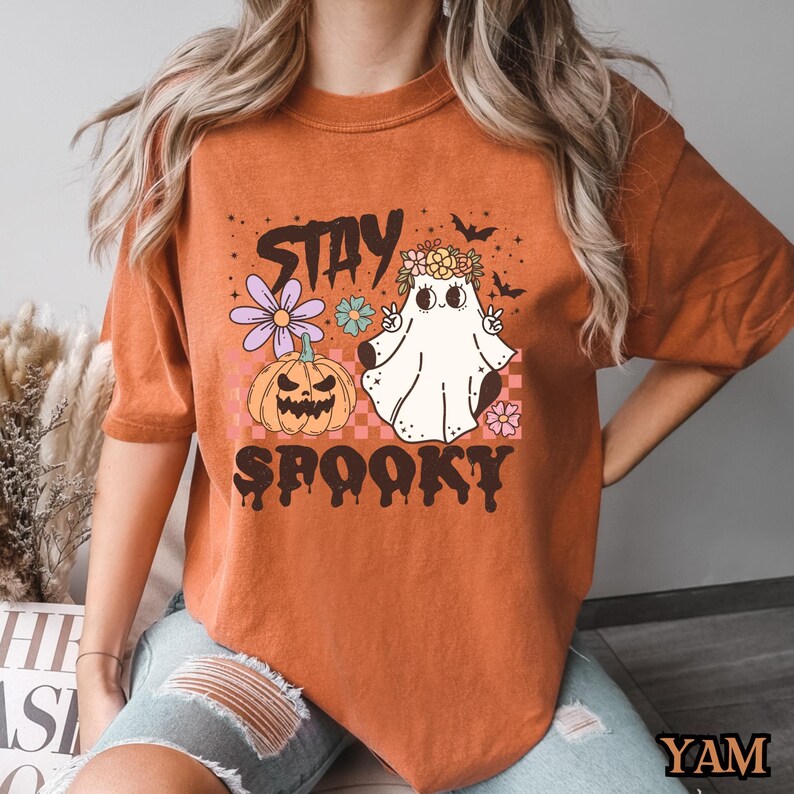 K&ouml;nnte beinhalten: Orangefarbenes T-Shirt mit einem Geist, der eine Blumenkrone tr&auml;gt und ein Peace-Zeichen macht. Der Text "Stay Spooky" ist mit einem K&uuml;rbis und Blumen auf das Shirt gedruckt.