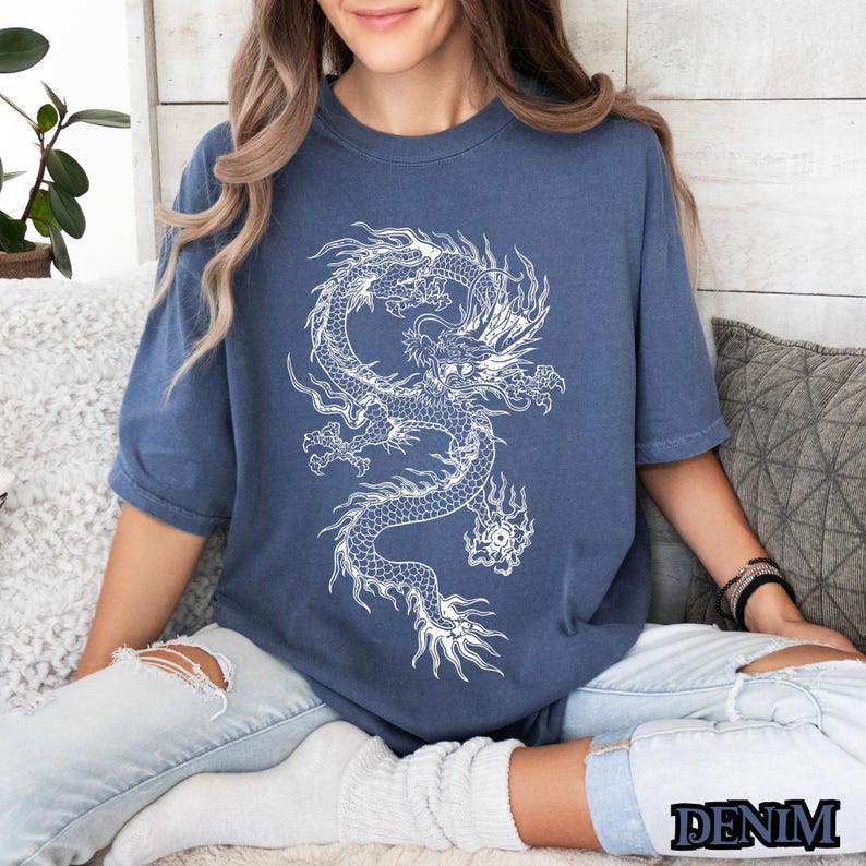 May include: A woman wearing a blue t-shirt with a white graphic of a dragon. The dragon is curled up in a circle with its head facing the viewer. The t-shirt is a relaxed fit and has a crew neck.