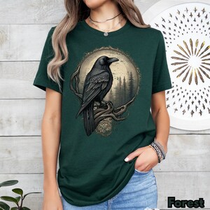 May include: A dark green t-shirt with a graphic of a black raven perched on a branch in a moonlit forest. The raven is detailed with a realistic design and the forest is depicted with a vintage aesthetic.