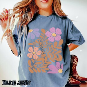 Op de afbeelding: Een blauw T-shirt met een bloemenprint met roze, oranje en paarse bloemen met bruine stelen en bladeren. De tekst "BLUE JEAN" is zichtbaar onderaan de afbeelding.