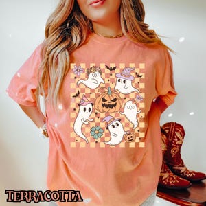 May include: A terracotta orange t-shirt with a graphic print of five cartoon ghosts and a pumpkin on a checkered background. The ghosts are wearing hats and have different expressions.