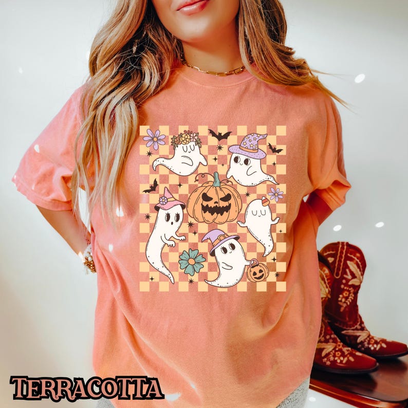 May include: A terracotta orange t-shirt with a graphic print of five cartoon ghosts and a pumpkin on a checkered background. The ghosts are wearing hats and have different expressions.