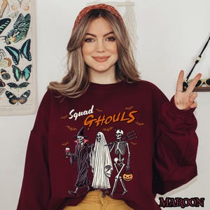 Pu&ograve; includere: Una felpa color granata con una grafica di tre scheletri vestiti da ghoul. Il testo "Squad Ghouls" &egrave; stampato sopra gli scheletri.
