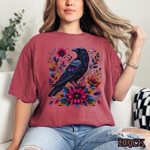 May include: A red t-shirt with a black crow surrounded by colorful flowers. The crow has blue and purple details on its wings and body.