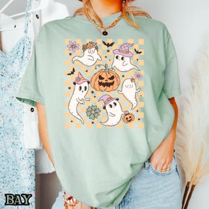 May include: A light green t-shirt with a cute Halloween design featuring five ghosts, a pumpkin, and a checkered pattern. The ghosts are wearing hats and have different expressions.