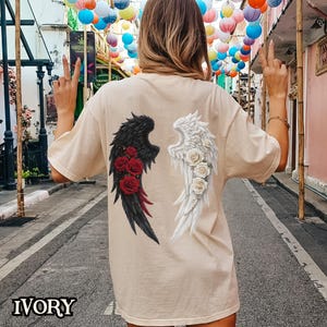 May include: A beige t-shirt with a graphic of black and white angel wings with red and white roses on each wing. The text "IVORY" is at the bottom of the image.