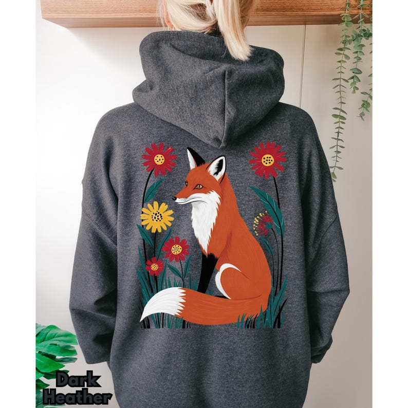 May include: Dark heather gray hoodie featuring a vibrant illustration of a fox amidst red and yellow flowers. The fox has orange fur, a white chest, and a fluffy white tail. The text "Dark Heather" is at the bottom of the hoodie.