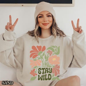 May include: Sand-coloured sweatshirt featuring a floral design in coral, peach, and green, with the words "STAY WILD" in green. The word "SAND" is printed in the bottom left corner. The sweatshirt is worn with a beige beanie.
