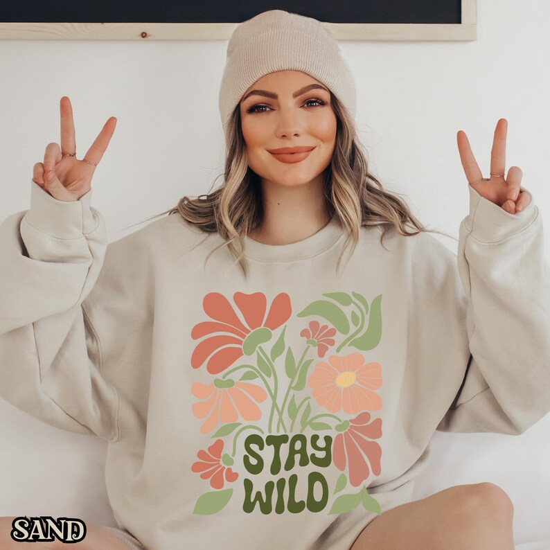 May include: Sand-coloured sweatshirt featuring a floral design in coral, peach, and green, with the words "STAY WILD" in green. The word "SAND" is printed in the bottom left corner. The sweatshirt is worn with a beige beanie.