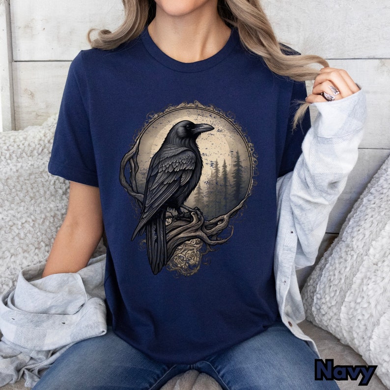 May include: A navy blue t-shirt with a graphic of a black raven perched on a branch in front of a moon and forest. The raven is in a circular frame with an ornate design.