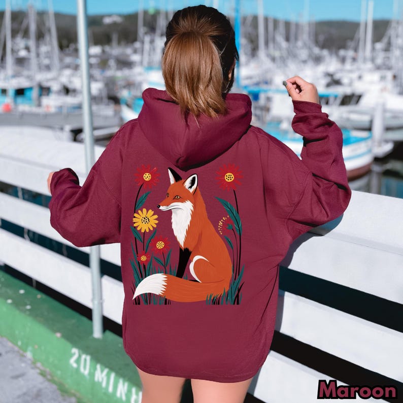 May include: Maroon hoodie featuring a vibrant illustration of a fox surrounded by flowers. The fox is depicted in shades of orange, white, and black, set against a backdrop of red and yellow flowers. The word "Maroon" is printed at the bottom right.