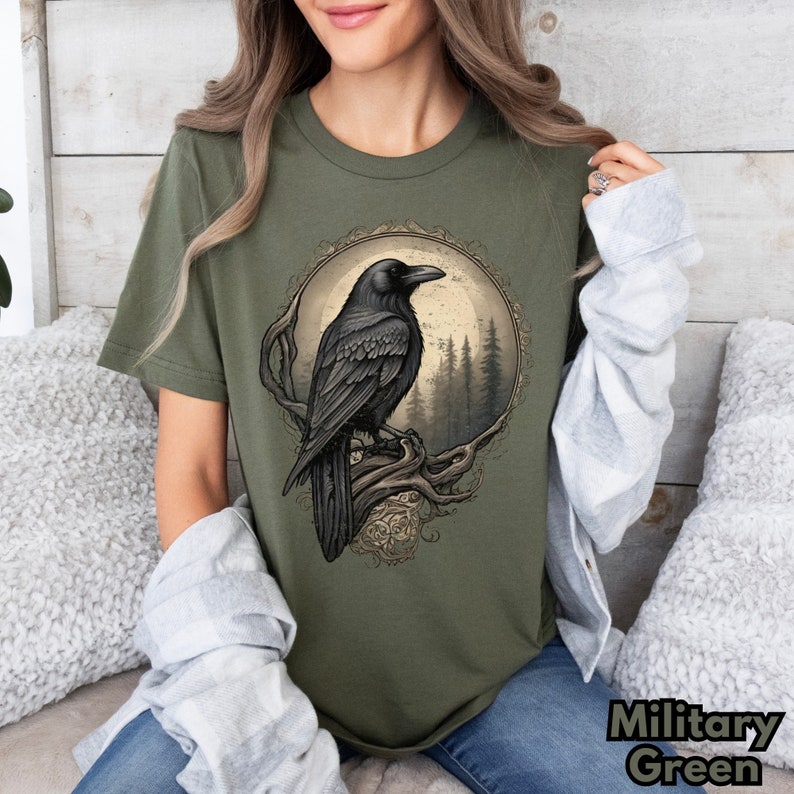 May include: A green t-shirt with a black raven illustration in a circular frame. The raven is perched on a branch with a full moon and forest in the background.