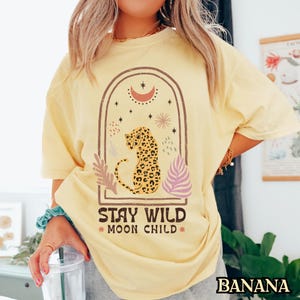 May include: A pale yellow t-shirt featuring a graphic print of a leopard inside an arched frame. The design includes a crescent moon, stars, and botanical elements with the text "STAY WILD MOON CHILD" printed in a dark brown, distressed font.