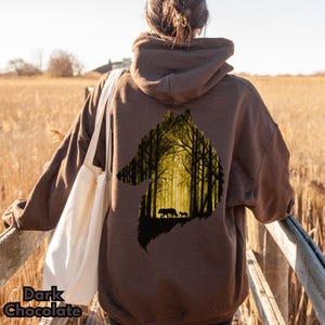May include: A dark brown hooded sweatshirt featuring a large graphic on the back. The graphic is shaped like a wolf head silhouette and contains a detailed scene of a forest with tall trees in yellow and black, with two wolves walking inside.