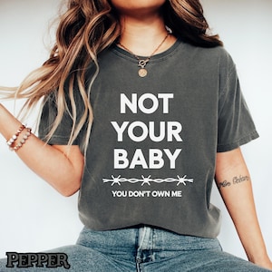 以下が含まれることがあります： 「NOT YOUR BABY」の文字が白の太字でプリントされた濃いグレーのTシャツ。テキストの下には有刺鉄線のデザインがあり、「YOU DON'T OWN ME」というフレーズがプリントされています。カジュアルなスタイルのTシャツです。