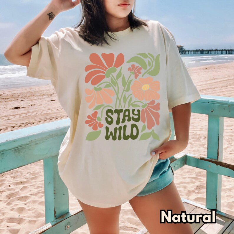 May include: A cream colored t-shirt with a floral design and the text "Stay Wild" in green letters. The shirt is being worn by a person on a beach.