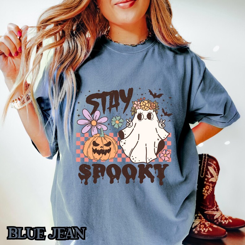 K&ouml;nnte beinhalten: Ein blaues T-Shirt mit einem Aufdruck eines Geistes, der eine Blumenkrone tr&auml;gt und ein Peace-Zeichen macht. Der Text "Stay Spooky" ist unter dem Geist gedruckt.
