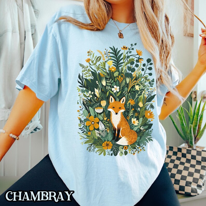 May include: A light blue t-shirt with a graphic of a fox surrounded by flowers and greenery. The fox is orange and white with a bushy tail.