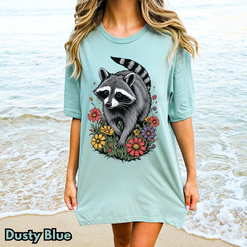 May include: A light blue t-shirt with a graphic of a raccoon surrounded by colorful flowers. The raccoon is gray and black with a white mask.