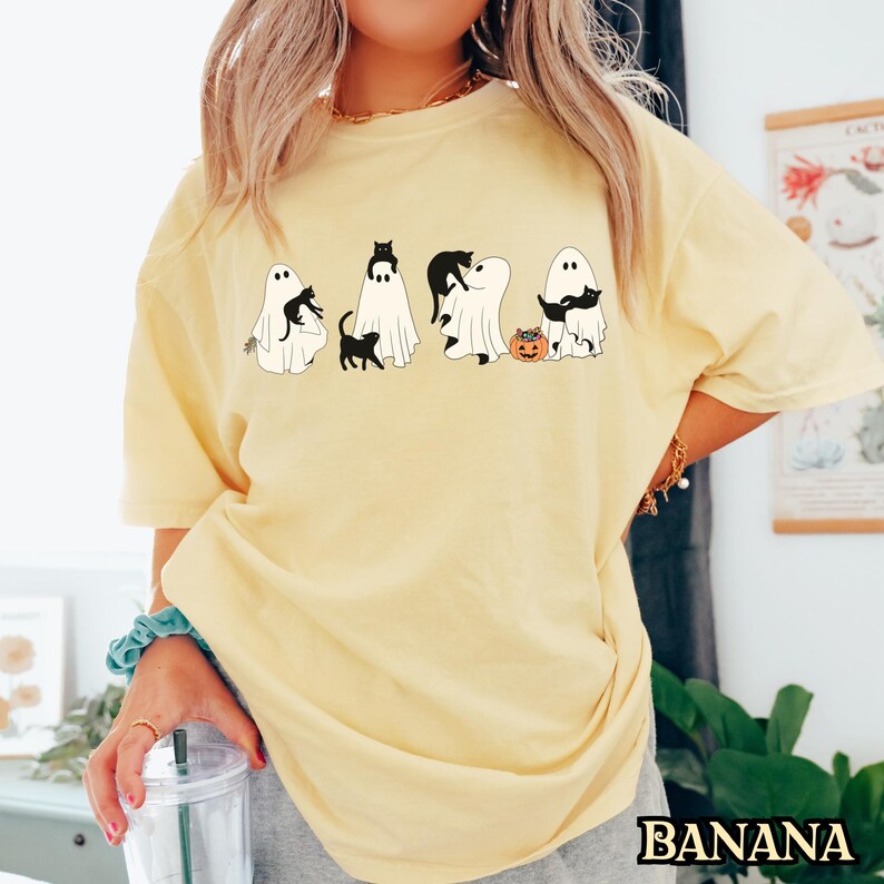 K&ouml;nnte beinhalten: Ein gelbes T-Shirt mit einem Aufdruck von schwarzen Katzen und Geistern. Der Aufdruck zeigt f&uuml;nf Geister mit schwarzen Katzen darauf. Die Geister halten K&uuml;rbisse und die Katzen tragen Hexenh&uuml;te. Der Text "BANANA" ist in der rechten unteren Ecke des Bildes sichtbar.