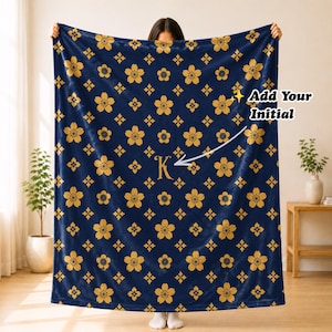May include: A navy blue fleece blanket featuring a repeating pattern of gold cherry blossoms and small diamond shapes. The center displays a large gold letter K, with text in the upper right corner that reads Add Your Initial.