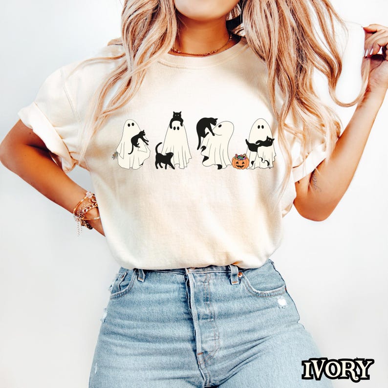 K&ouml;nnte beinhalten: Ein wei&szlig;es T-Shirt mit einem Aufdruck von f&uuml;nf Geistern mit schwarzen Katzen darin. Die Geister tragen Halloween-Kost&uuml;me und halten K&uuml;rbisse. Das T-Shirt wird von einem Model getragen, das eine blaue Jeans tr&auml;gt.