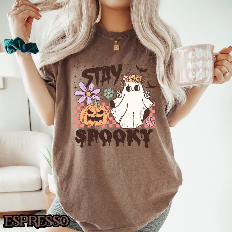 K&ouml;nnte beinhalten: Ein braunes T-Shirt mit dem Text "Stay Spooky" in einer tropfenden Schriftart. Das Shirt zeigt einen Cartoon-Geist mit einer Blumenkrone und einem K&uuml;rbis.