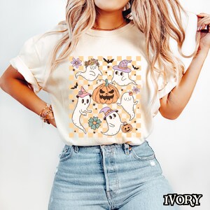 May include: A white t-shirt with a graphic of five cartoon ghosts, a pumpkin, and a checkered pattern. The ghosts are wearing hats and flowers. The background is a light orange color.