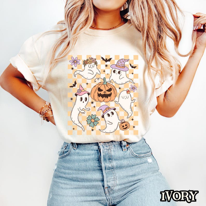 May include: A white t-shirt with a graphic of five cartoon ghosts, a pumpkin, and a checkered pattern. The ghosts are wearing hats and flowers. The background is a light orange color.