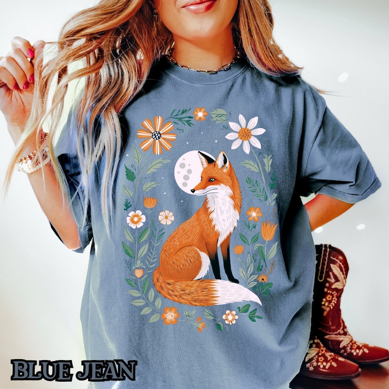 Op de afbeelding: Een blauw T-shirt met een afbeelding van een vos die in een bloemenveld onder een volle maan zit. De vos is oranje en wit, en de bloemen zijn in verschillende kleuren.