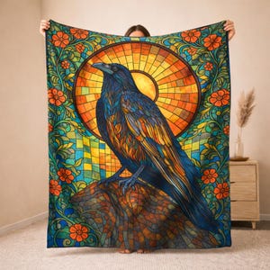 Peut inclure: Une grande couverture colorée avec un motif de style vitrail. Le motif comprend un corbeau détaillé perché sur un rocher, sur fond de soleil et d'éléments floraux dans des tons de bleu, d'orange et de vert.