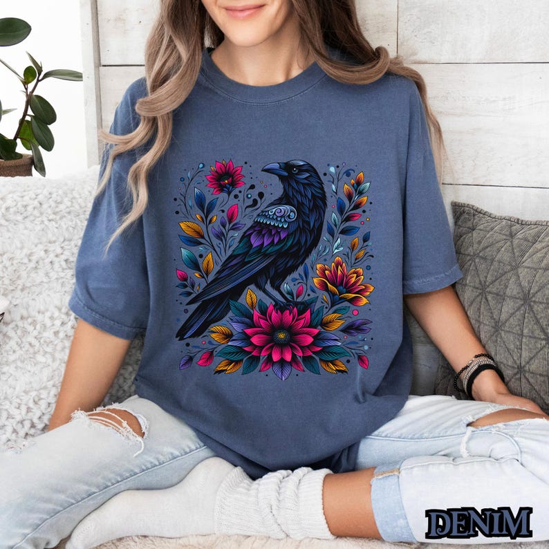 May include: A dark blue t-shirt with a black crow surrounded by colorful flowers. The crow has intricate details and the flowers are a mix of pink, purple, and yellow.